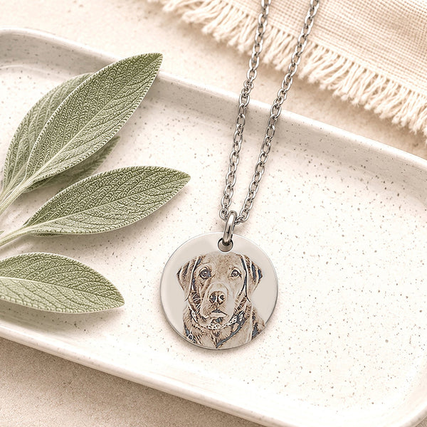 Personalised pet portrait necklace displayed on a ceramic trinket tray with sage leaves, showcasing a custom engraved dog portrait on a stainless steel pendant.